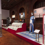 Wood-paneled, interior exhibition space with multiple examples of personal, protective equipment, including face masks, face-shields and hospital scrubs, displayed on grey standing mannequins, grey mannequin heads and in colorful posters and blown-up photographs.