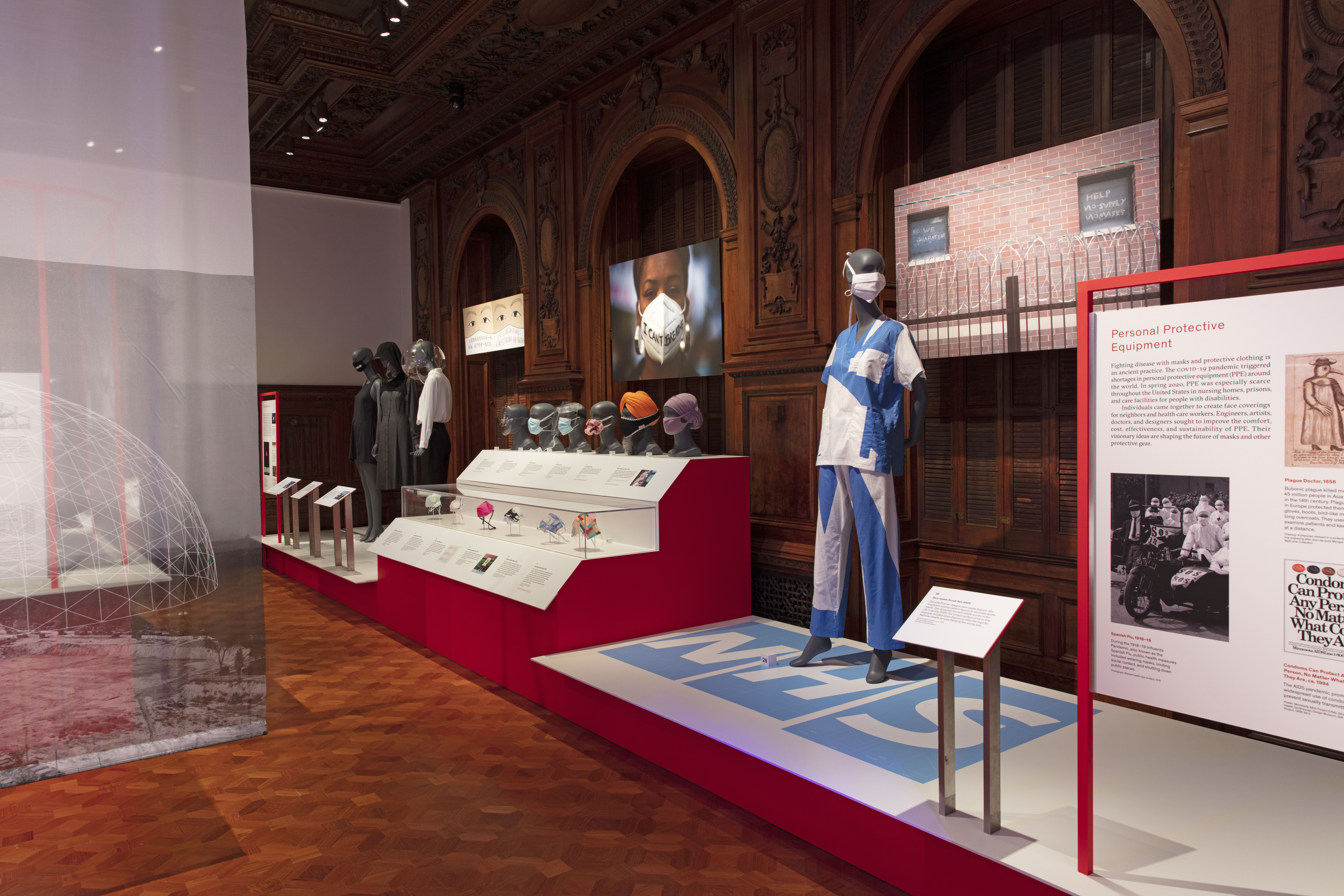 Wood-paneled, interior exhibition space with multiple examples of personal, protective equipment, including face masks, face-shields and hospital scrubs, displayed on grey standing mannequins, grey mannequin heads and in colorful posters and blown-up photographs.