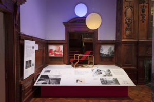 A red-brown armchair with a ribbon-like seat stands side-on on a plinth whose surface is white and filled with photographs and explanatory text. Two orb-like yellow and blue lights hang above, behind are two photographs on either side of a wood-paneled doorway