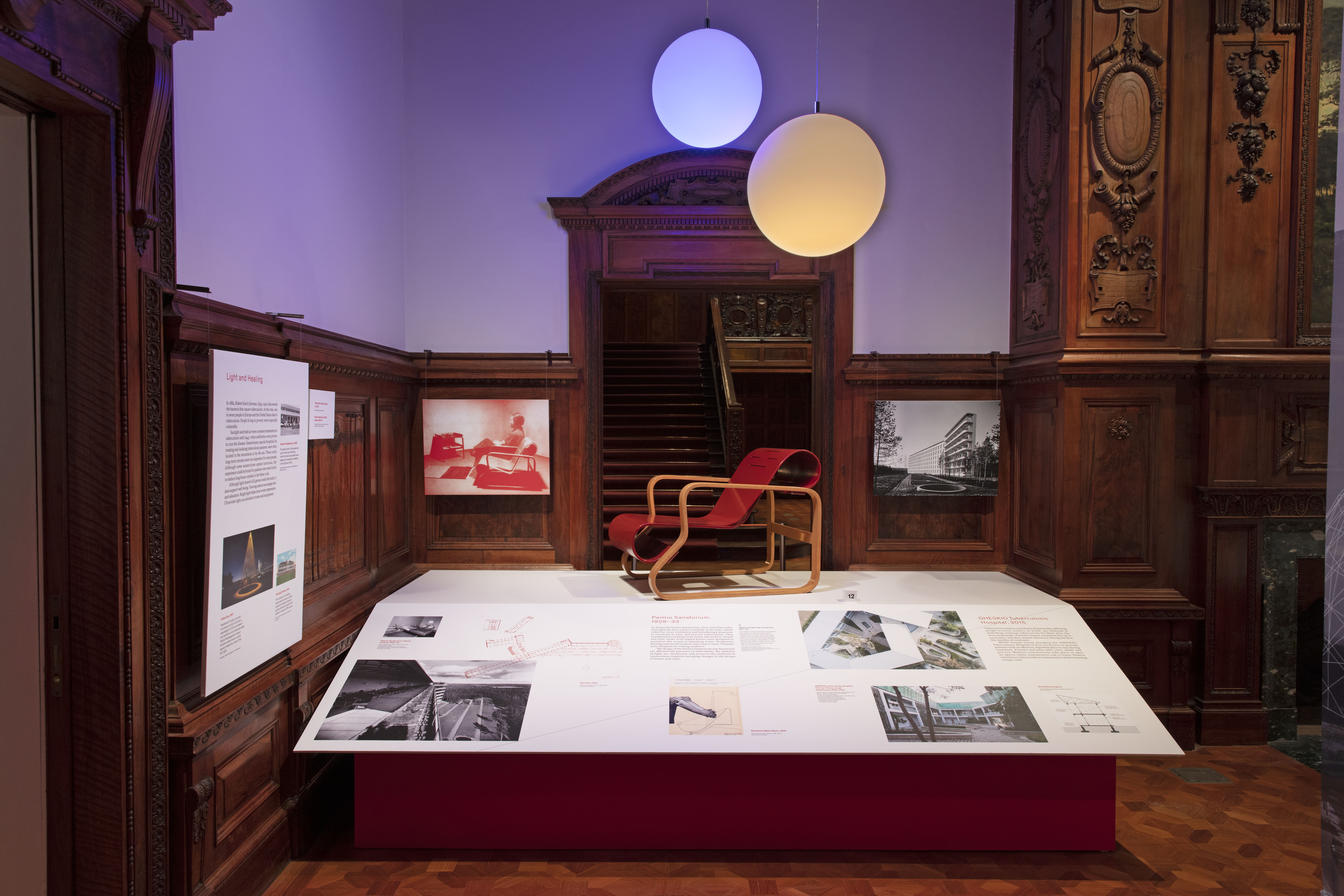A red-brown armchair with a ribbon-like seat stands side-on on a plinth whose surface is white and filled with photographs and explanatory text. Two orb-like yellow and blue lights hang above, behind are two photographs on either side of a wood-paneled doorway