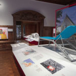 Wood-paneled interior exhibition space with posters, photographs, objects, and panels displayed; most prominent is a blue, reclining chair that stands on a plinth, and a photograph showing the exterior of a cholera treatment center.