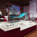 Exhibition space with a large white central pedestal displaying a reclined teal blue cot and a small white and blue building model. In the left background are two mannequins, one wearing a black hood and the other a clear plastic bubble helmet.