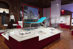 Exhibition space with a large white central pedestal displaying a reclined teal blue cot and a small white and blue building model. In the left background are two mannequins, one wearing a black hood and the other a clear plastic bubble helmet.