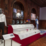 Wood-paneled, interior exhibition space displaying examples of personal, protective equipment, including multi-colored face masks, transparent face-shields, and blue and white hospital scrubs, displayed on grey standing mannequins, grey mannequin heads and in colorful posters and blown-up photographs.