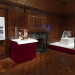 Wood-paneled exhibition space with two display platforms split by a fireplace, on the left is a geometric model of a hospital encased in plexiglass, on the right is a clear plastic hood with draping sheeting on the back.