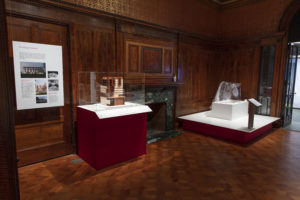 Wood-paneled exhibition space with two display platforms split by a fireplace, on the left is a geometric model of a hospital encased in plexiglass, on the right is a clear plastic hood with draping sheeting on the back.