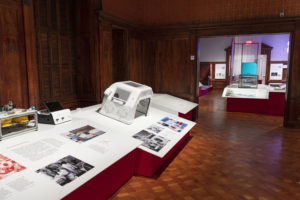 Exhibition space with a white angle-fronted pedestal along the left wall, printed with colorful graphics and text, displaying ventilation devices. A large doorway on the back wall opens into another gallery.