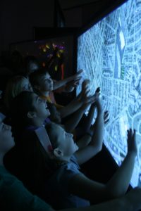 A photograph of several children smiling as they play with an interactive map on a screen.