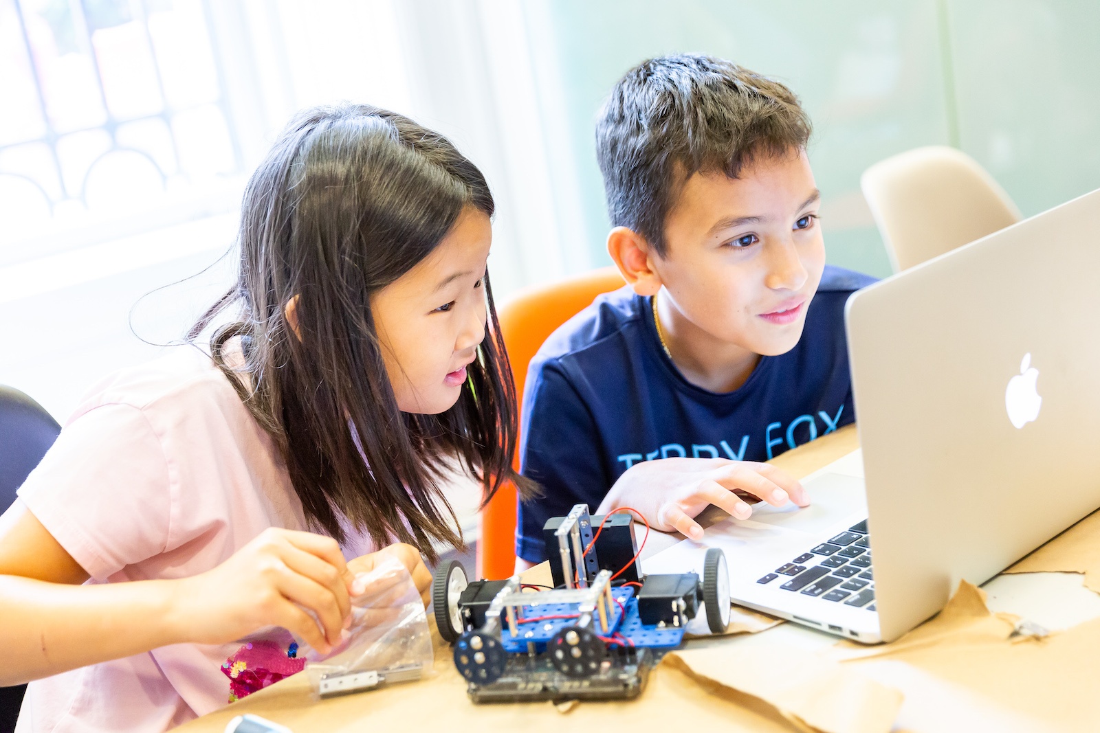 Two children sit behind a desk looking at a laptop screen, one, on the right, is medium-skinned with shoulder-length black hair, the other is medium-skinned with short dark hair, in the foreground is a wheeled device with multi-colored wires.