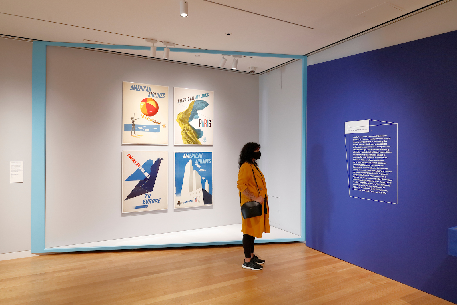 A person with curly black hair dressed in a yellow coat stands in an exhibition space reading white text on a royal blue wall with a wall of four, brightly-colored, graphic posters advertising American airlines behind