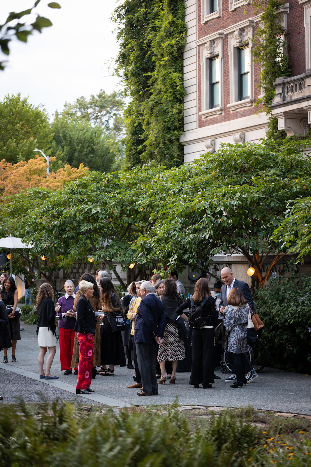 A group of well-dressed people standing drinking and talking in the Cooper Hewitt garden. There is a line of green trees behind them and part of the back façade of the Georgian-style mansion