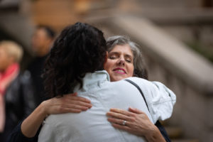 Two people embracing, one, their back to us, wears a white shirt and has curly, dark hair, the other, facing us, is light-skinned with wavy, grey hair, photographed with a blurred background