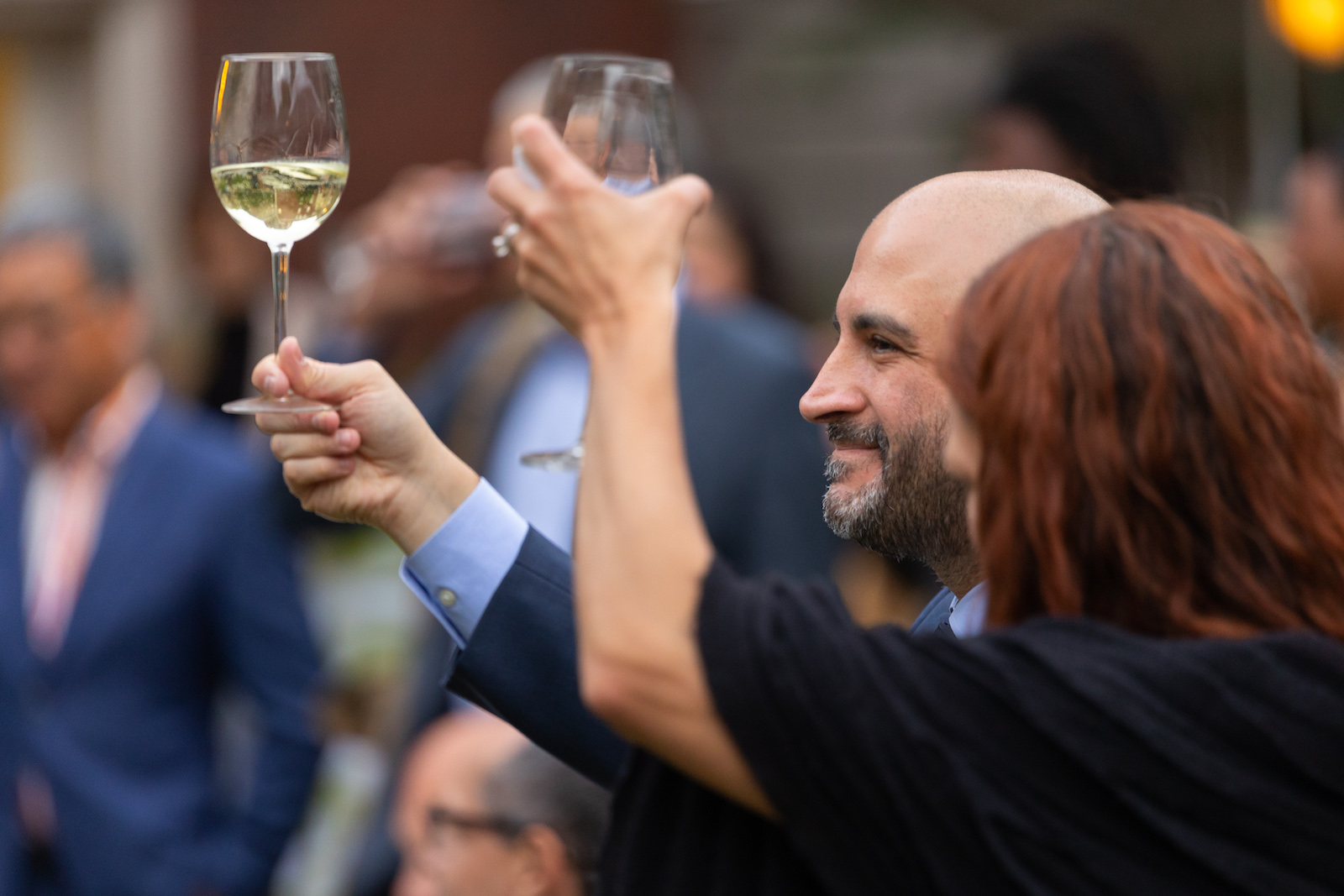 Two people raise glasses in celebration, one, turned away from us, has wavy red-brown hair and wears black, the other is light-skinned, bald with a cropped beard and wears a navy-blue suit