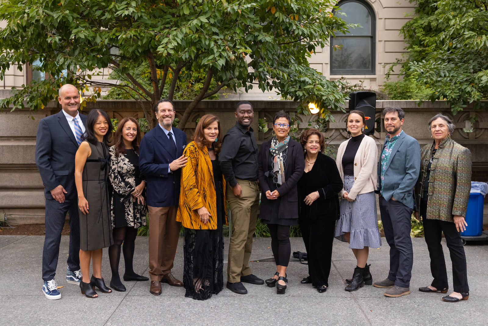 A group of well-dressed people stand in a line in the Cooper Hewitt garden, they are posed in front of a grey stone wall and a green tree