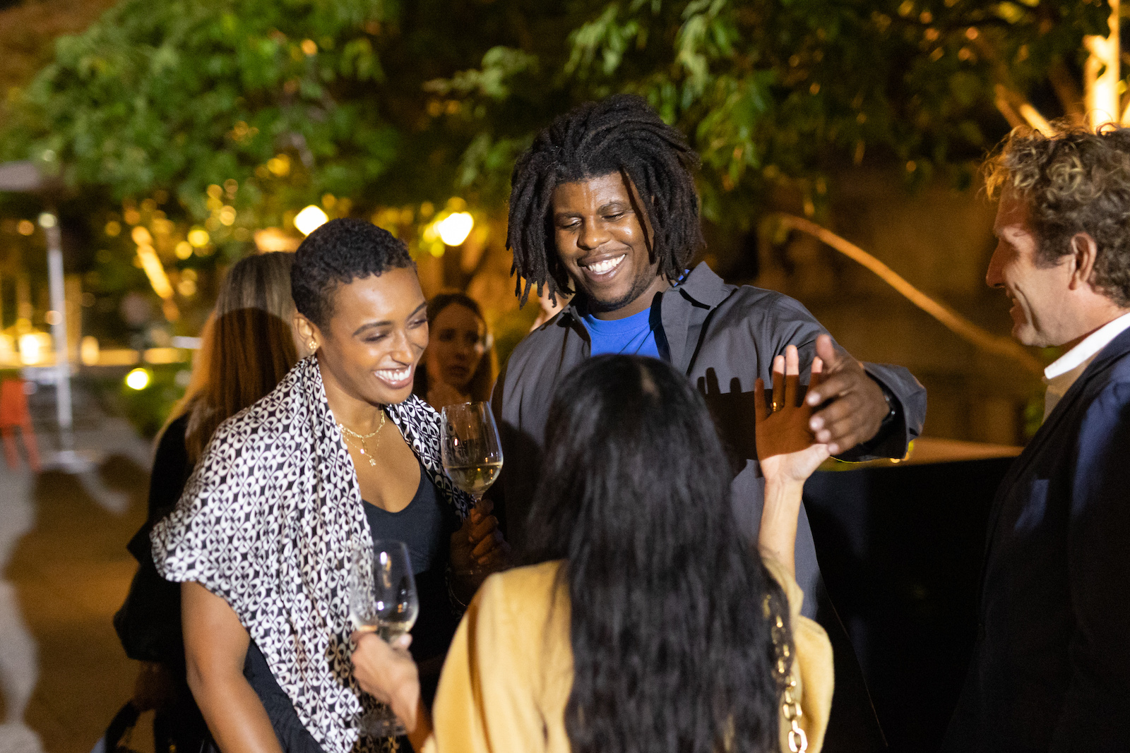 A group of four people stand in a circle smiling holding wine glasses, one in the center, their back to us, wears yellow, to their left is a medium-skinned woman, to her left is dark-skinned man, on the right is a light-skinned man