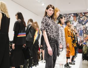 A photograph of a dozen models on a runway arranged in two lines shoulder-to-shoulder. Each model has a different outfit, the model closest to the camera is looking directly at the camera and is wearing a black blouse with white flower pattern and white and black striped pants.