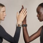Two models facing each other in profile with their right arms raised and their hands touching in the center. The model on the left wears a white cuff on her wrist and the model on the right wears a gold cuff. The model on the left is a blonde white woman in a dark grey turtleneck sweater. The model on the right is a Black woman with short black hair in a sleeveless black turtleneck with a turquoise zipper.