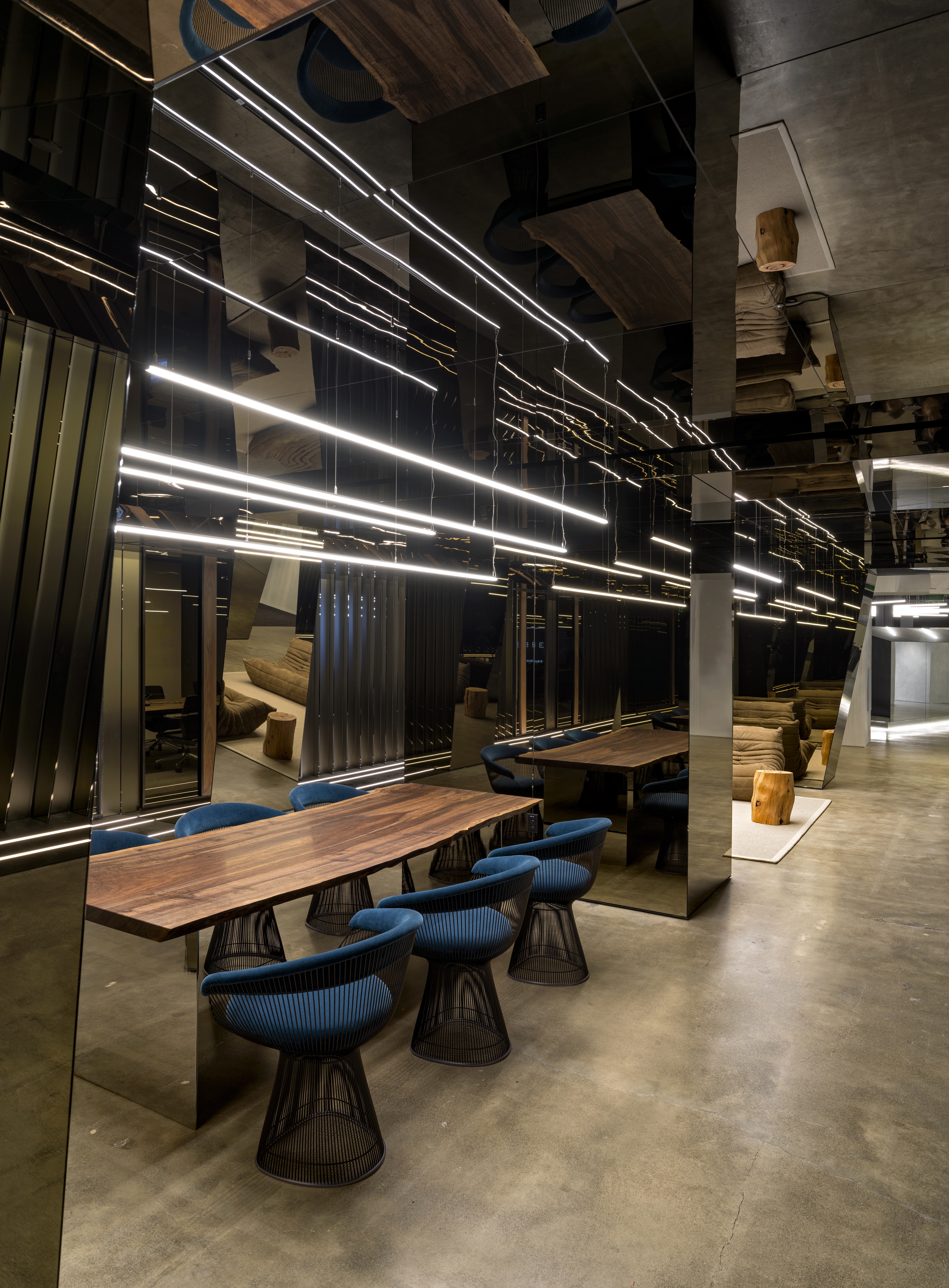 A photograph of the Uber headquarters with long LED lights hanging above a wooden table. The lights reflect off of the dark mirrored walls. The table has size low back dark turquoise chairs around it.