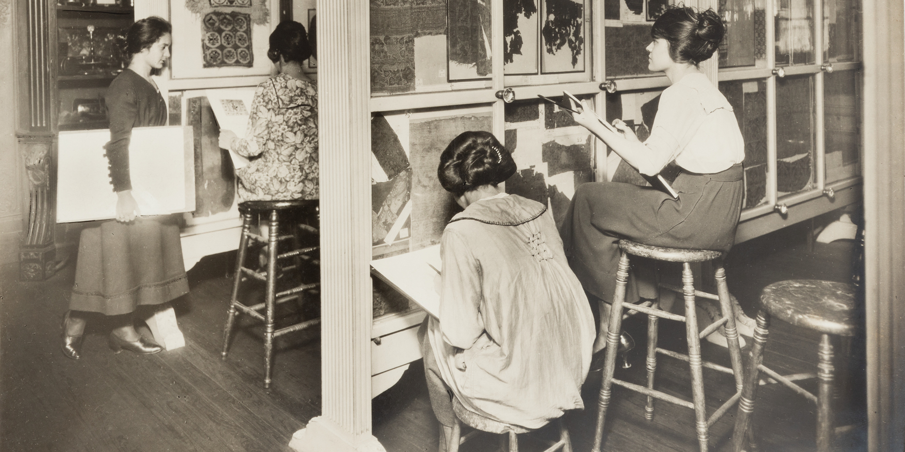 Black and white photograph of a group of students in a room studying different styles of textiles.