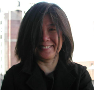A photograph of Yeohlee Teng smiling at the camera with her hair covering the left half of her face.