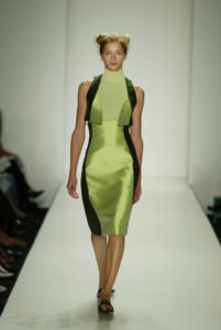 A photograph of a model walking down a runway in a green, knee-length dress and black sandals. The sides of the dress are black.