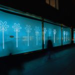 A photograph taken at night of a store window display. The background of the display is blue with four white LED flower shapes. There is text on the window but all that can be read is the title [Flower Power].