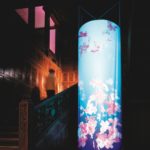 An installation in Cooper Hewitt's Great Hall. The cylindrical shaped structure emits light from within and has pink cherry blossoms adorning the blue structure.