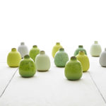 A photograph of thirteen ceramic jugs of the same shape and size. The jugs are varying shades of green and white and are arranged on a white surface with a white background.