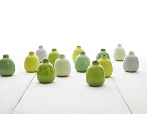 A photograph of thirteen ceramic jugs of the same shape and size. The jugs are varying shades of green and white and are arranged on a white surface with a white background.