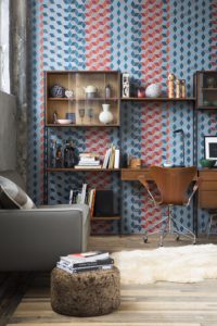A photograph of a room with a chair and coffee table with books on it to the left. Against the tile patterned blue and red back wall is a desk and shelves with books, ceramics, bottles, lamps, and sculptures displayed. On the wooden floors is a small white fur rug.