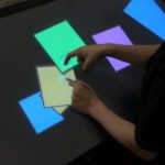A photograph taken from above of a person using an interactive touch table using one finger on their right hand and using a stylus in their left hand. On the screen are blue, yellow, green, and purple squares.