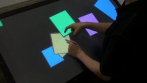 A photograph taken from above of a person using an interactive touch table using one finger on their right hand and using a stylus in their left hand. On the screen are blue, yellow, green, and purple squares.