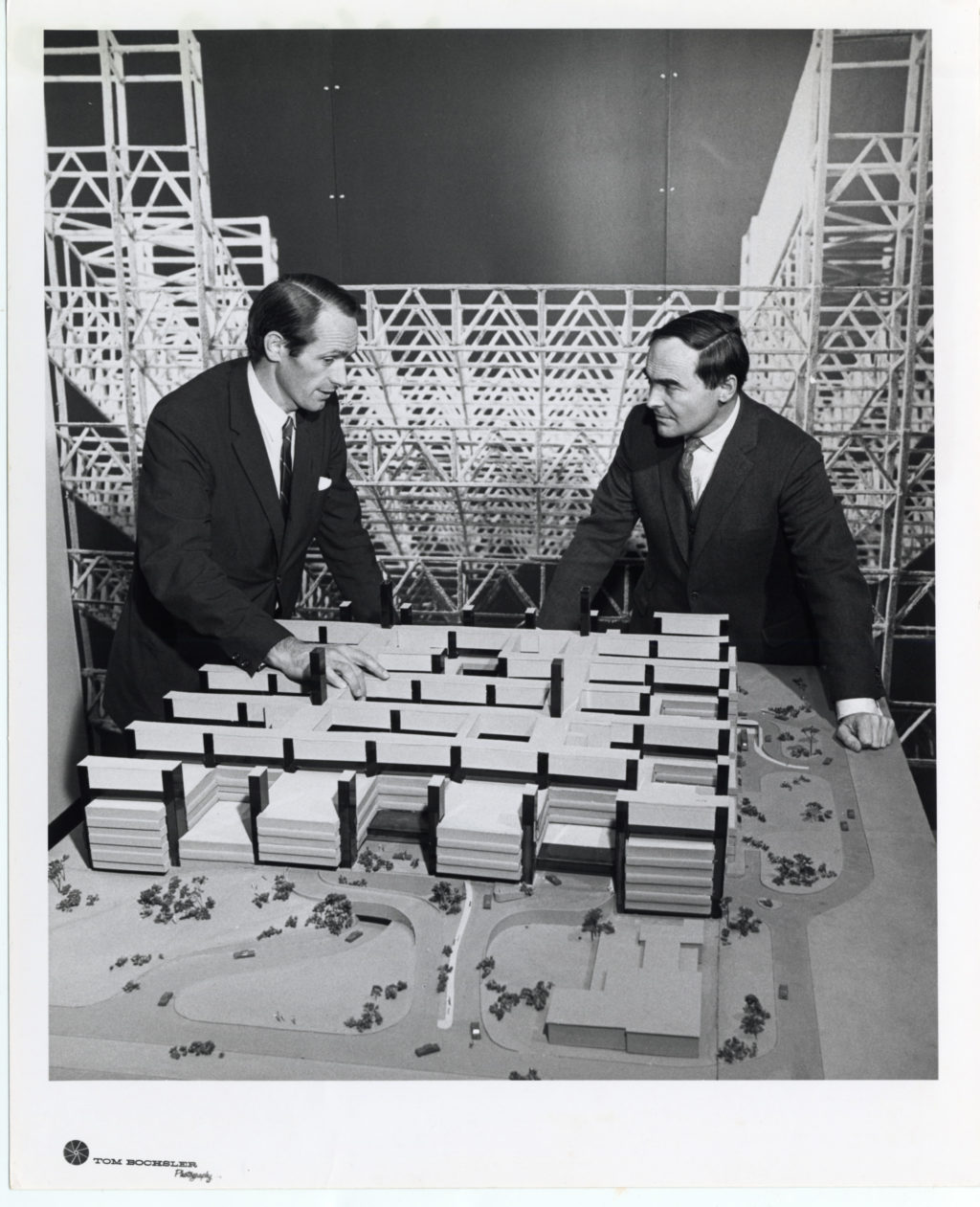 Black and white photograph showing two men in suits standing in front of a large architectural model made up of a grid of squares. Behind them, rows of scaffolding.