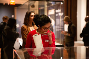 In the foreground, a South Asian woman looks down into a glass case, the contents of which are not visible. She is wearing a red sweater and dark rimmed glasses, touching her face with her left index finger pensively. In her right hand, she's holding a folded worksheet. Behind her scattered around are other individuals looking at objects and holding worksheets.