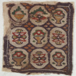 A chocolate brown square fragment of a woven wool and linen tapestry with nine circular cream sections containing stylized colorful flowers and bowls of fruit split by a repeating ochre pattern.