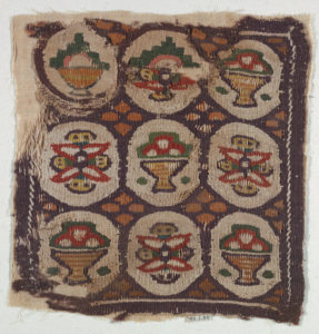 A chocolate brown square fragment of a woven wool and linen tapestry with nine circular cream sections containing stylized colorful flowers and bowls of fruit split by a repeating ochre pattern.