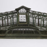 A birdcage in the shape of the Rialto bridge in Venice, with a wooden frame painted in olive green and intricate wire scrollwork along the sides, door and top.