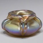 Free-blown glass vase with pale blue, purple, and gold-toned iridescent body, its shape is a depressed sphere with a squarish opening that has a turned-down rim. Three irregular thick ribs line the sides diminishing to points at the base.