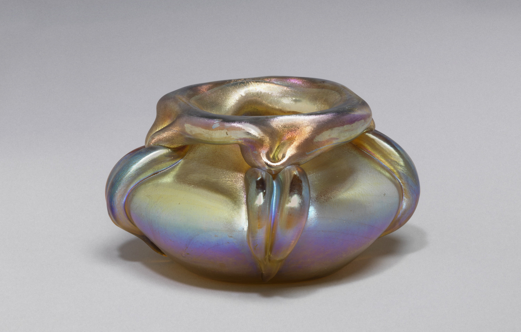 Free-blown glass vase with pale blue, purple, and gold-toned iridescent body, its shape is a depressed sphere with a squarish opening that has a turned-down rim. Three irregular thick ribs line the sides diminishing to points at the base.