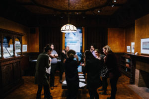 A group of people stand in the circle of a semi-darkened period room. At the periphery are graphics and paintings on display. The people are leaning towards each other, bending into the center of the circle. They are looking at pieces of paper sitting on a bench between them.