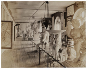 Faded photograph of a gallery space that has rows of chairs and sculptures of cherubs and urns on the right, on the left is a paneled storage wall with a panel containing two fans pulled out.