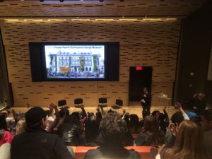 A view from behind of an audience in bleacher style seats in a blonde-wood-paneled room. On the far wall is a large screen that shows a picture of the facade of Cooper Hewitt, Smithsonian Design Museum. A woman is holding a microphone and some audiences members have their hands raised.