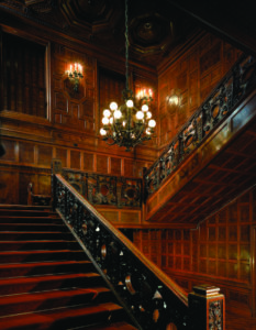 The image is a photograph of staircase. It rises on the left, turns ninety degrees to the right as a landing, then turns ninety degress again to continue ascending up the right. A round chandelier with about twenty round bulbs hangs in the center. The staircase and its surroundings are constructed of rich brown wood, and the staircase is carpeted in a deep red textile.