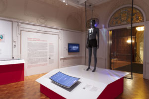 Exhibition space with grey standing mannequin on a plinth in center-left with figural projection on a transparent screen behind, a curved green screen in the foreground and a panel of explanatory text on the white back wall