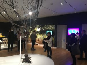A museum gallery with a dead, bare tree on display in the foreground. In the background, people are scattered around the room looking at objects on display and making notes. To the left of the far wall is a large image of what looks like a massive tree canopy. A screen with pink and blue light is visible off to the right.
