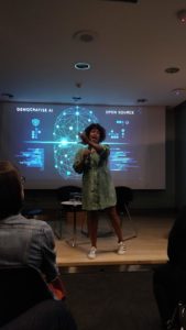 A light skinned Black woman wearing a green dress and white sneakers is speaking into a microphone while gesturing. She is standing on stage with a projected slide behind her. The slide depicts a glowing blue illustrations that reference artificial intelligence with a techy aesthetic. In the foreground to the left and right are seated audience members.