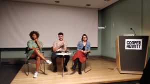 Three people are seated on a stage with small table in front of them. To their left is a large podium with a sign that reads Cooper Hewitt. Each of them looks expectantly in the direction of the camera with various expressions ranging from attentive to amused.