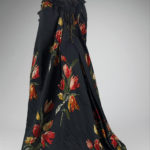 Rear view of a floor-length black silk evening cloak embroidered with a repeating pattern of large red and yellow parrot tulips with a sweeping train and a black lace collar.
