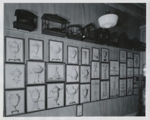 Photograph of interior with drawings of vessels on the wall, intricate birdcages sit atop a ledge above the drawings in a gallery.