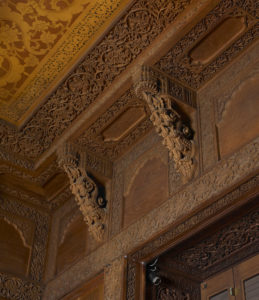 Photograph of a corner where two walls and a ceiling made of wood meet. Intricate designs are carved into the wood mimicking organic vine forms creating swirls of highly detailed lattice forms. Two wooden buttresses add additional ornamentation resembling ornate tentacles.